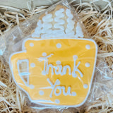 Decorative cookie shaped like a mug with 'Thank You' text, wrapped in clear plastic on straw.