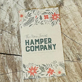 Floral design card with 'The New Forest Hamper Company' text on a wooden surface