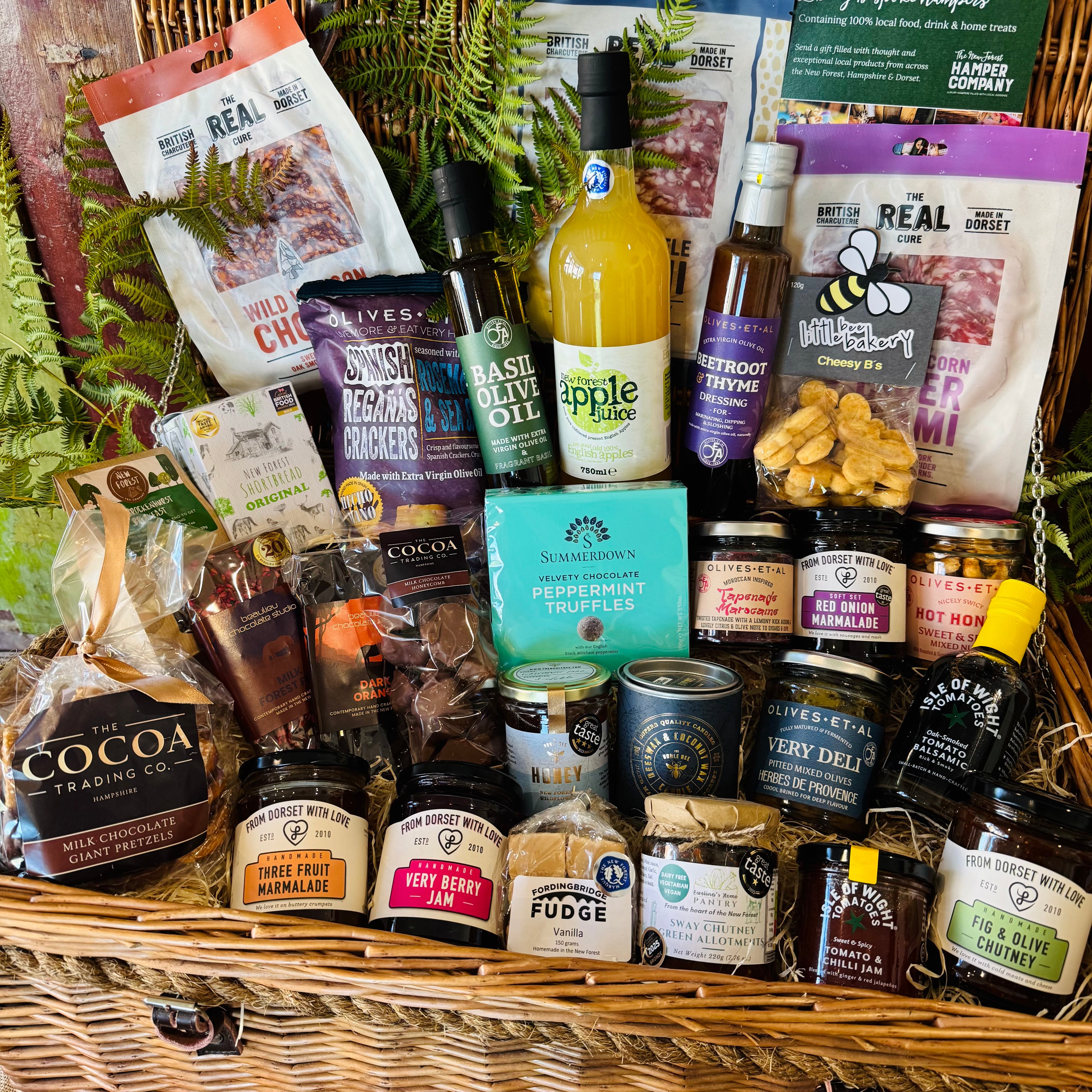 The Ultimate Hampshire & Dorset Goodies Hamper - a 24 inch lidded willow basket filled with Real Cure charcuterie, cheese biscuits, olives, local preserves, Beaulieu chocolate, New Forest apple juice and a Noble Bee beeswax candle