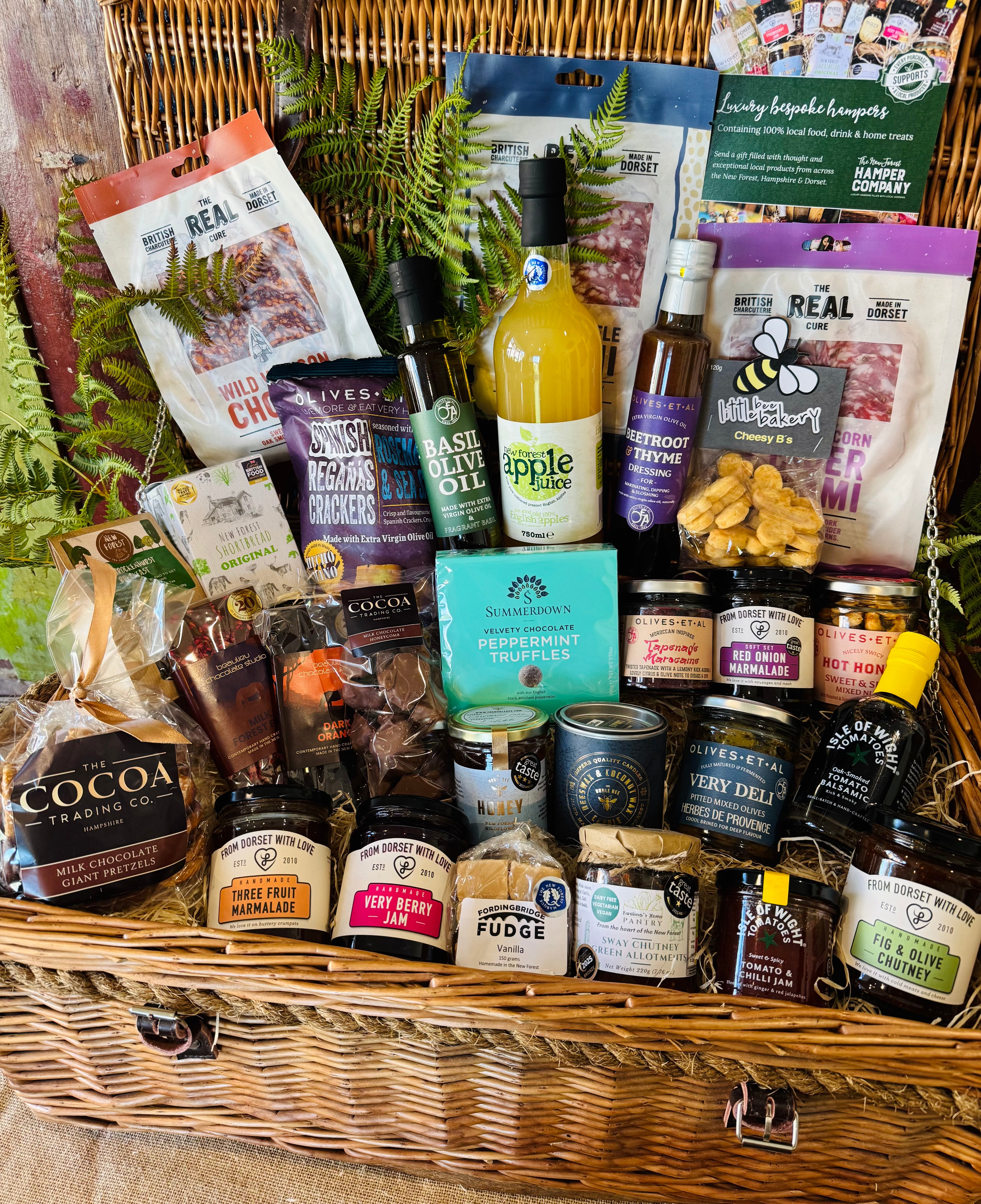 The Ultimate Hampshire & Dorset Goodies Hamper - a 24 inch lidded willow basket filled with Real Cure charcuterie, cheese biscuits, olives, local preserves, Beaulieu chocolate, New Forest apple juice and a Noble Bee beeswax candle
