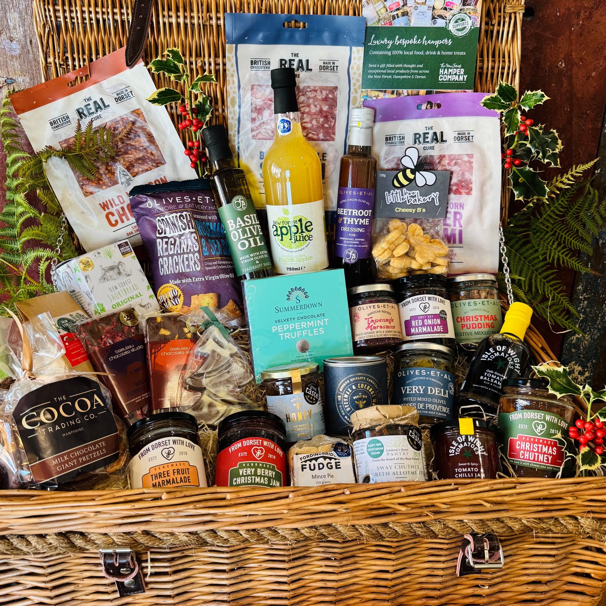 The Ultimate Christmas Goodies Hamper - a 24 inch lidded willow basket filled with Real Cure charcuterie, Cheesy Stars, festive preserves, chocolates, mince pie fudge, New Forest apple juice and a Christmas spice candle, dressed with holly and fern