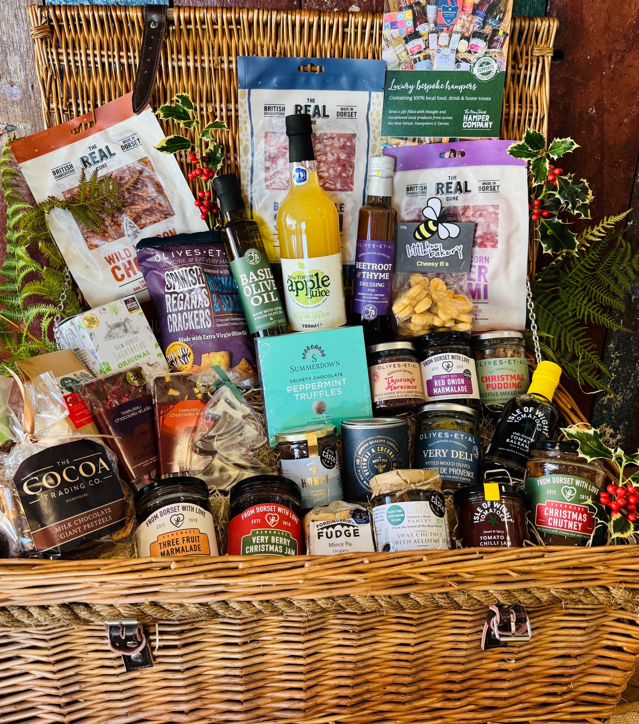The Ultimate Christmas Goodies Hamper - a 24 inch lidded willow basket filled with Real Cure charcuterie, Cheesy Stars, festive preserves, chocolates, mince pie fudge, New Forest apple juice and a Christmas spice candle, dressed with holly and fern