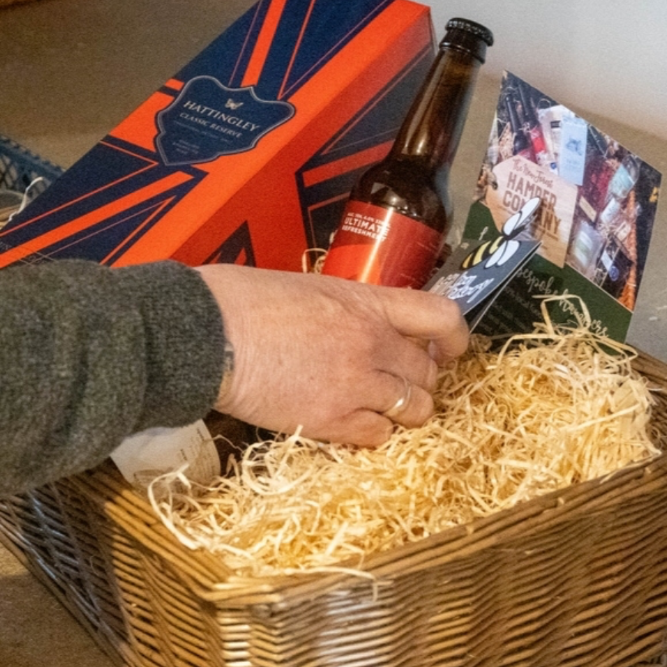 Person reaching into a gift basket with a beer bottle and cards, featuring a Union Jack box in the background.