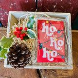 Little Bee Bakery hand-iced Ho Ho Ho Christmas biscuit in festive red with white lettering and a string of coloured Christmas lights, in a wooden gift crate with holly, red berries and a pinecone