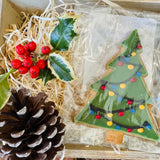 Little Bee Bakery hand-iced Christmas Tree biscuit decorated with garlands and red, blue and yellow baubles, in a wooden gift crate with holly, red berries and a pinecone