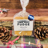 A 150g bag of Fordingbridge Fudge Mince Pie, wrapped in clear cellophane with a white label, displayed on a rustic wooden surface with pinecones, tinsel, and a plaid blanket.