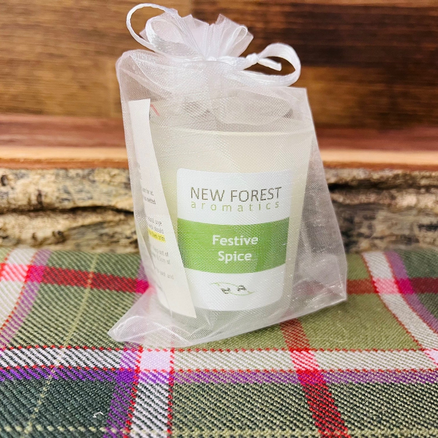 A white candle in a clear mesh gift bag with a white ribbon, labeled 'NEW FOREST aromatics Festive Spice', placed on a green and red plaid fabric surface with a rustic wood background.