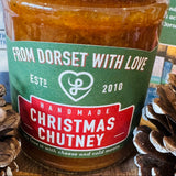 A close-up of a glass jar of Christmas Chutney with a black lid and a green and red label reading 'From Dorset With Love' and 'Handmade Christmas Chutney.' Pinecones sit on both sides on a wooden surface.