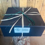 Blue box with white and green ribbon on a wooden surface, featuring 'Hamper Company' label.