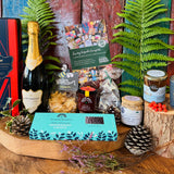 The Hattingley Christmas Luxury Hamper laid out - Hattingley Classic Reserve English sparkling wine in its red and navy giftbox, Noble Bee wildflower honey and Christmas Spice candle, Summerdown peppermint crisps, Beaulieu chocolate Christmas tree, Ewelina's spiced plum jam, Isle of Wight tomato and chilli jam and Little Bee cheesy stars, on an oak board with ferns and holly
