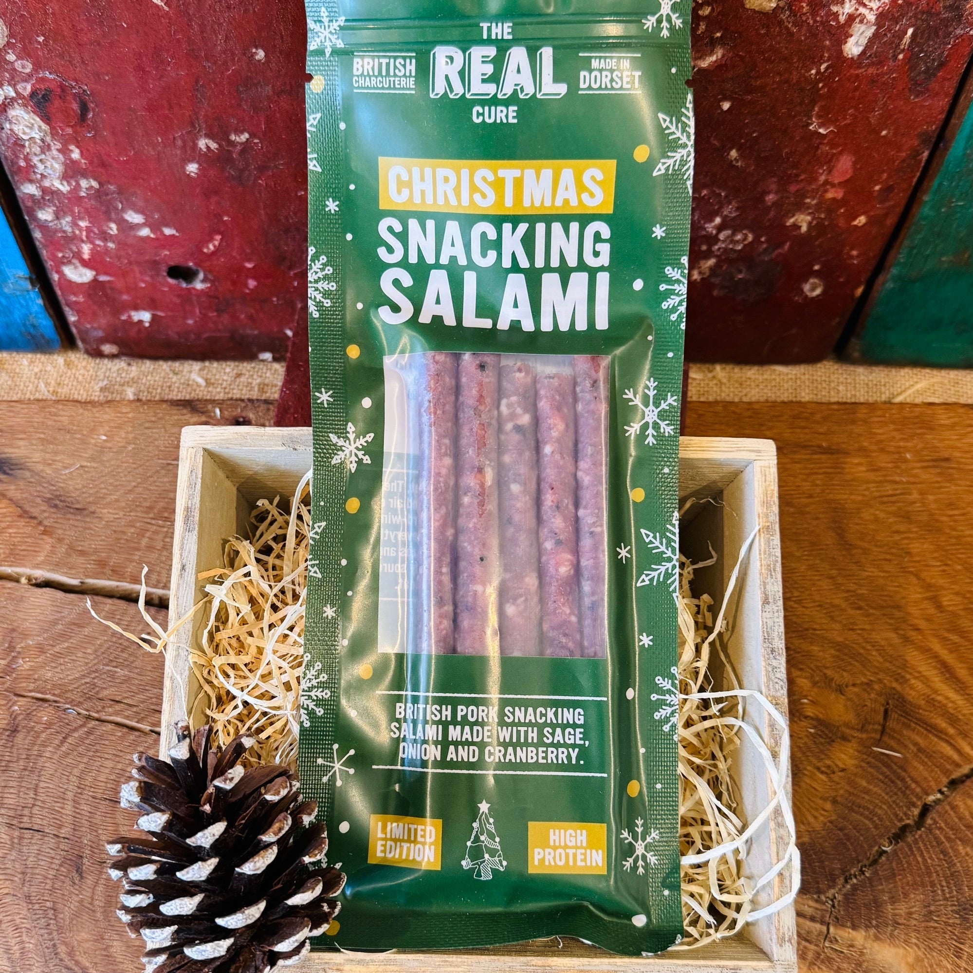 The Real Cure Christmas Snacking Salami, a green limited edition pack of five British pork sticks made with sage, onion and cranberry, in a wooden gift crate with wood wool and a pinecone