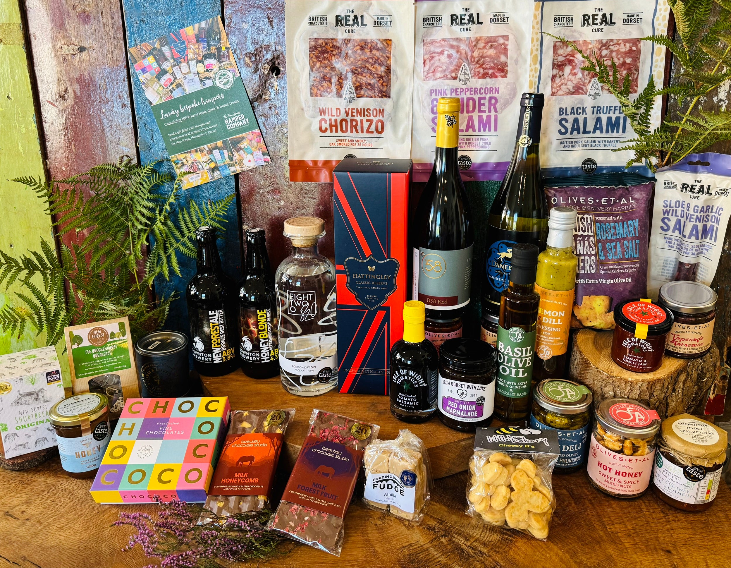 The Ultimate Hampshire & Dorset Hamper shown in display format - Hattingley Valley Classic Reserve English sparkling wine, B58 red, Danebury Madeleine Angevine white, 820 Spirits London Dry Gin and two Downton Brewery ales with Real Cure charcuterie, Chococo chocolates, Olives et Al oils, olives and preserves and the 24 inch lidded willow basket