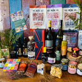 Full contents of The Ultimate Hampshire & Dorset Hamper laid out - six Hampshire bottles, four Real Cure salamis and chorizo, Isle of Wight Tomatoes balsamic and chilli jam, From Dorset With Love red onion marmalade, Sway Chutney, New Forest wildflower honey, Chococo and Beaulieu chocolates, shortbread, fudge and a Noble Bee beeswax candle