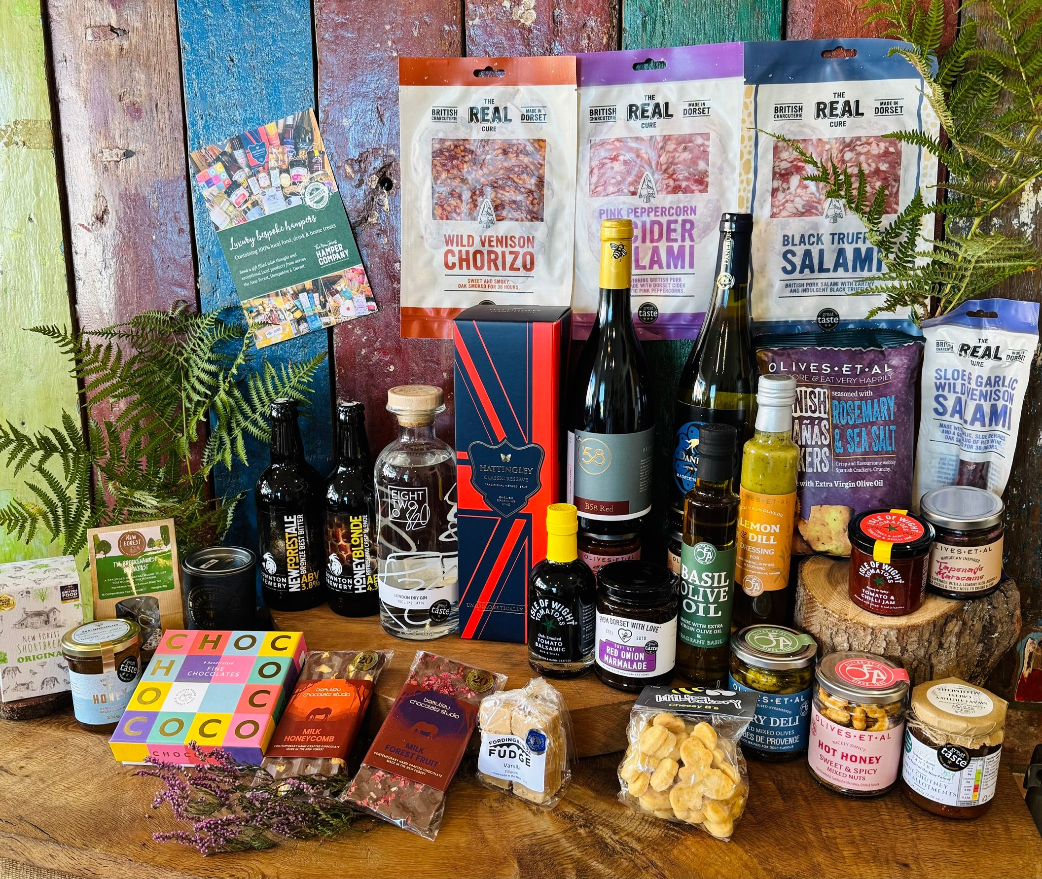 Full contents of The Ultimate Hampshire & Dorset Hamper laid out - six Hampshire bottles, four Real Cure salamis and chorizo, Isle of Wight Tomatoes balsamic and chilli jam, From Dorset With Love red onion marmalade, Sway Chutney, New Forest wildflower honey, Chococo and Beaulieu chocolates, shortbread, fudge and a Noble Bee beeswax candle